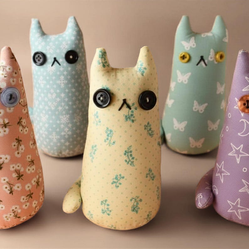May include: Five handmade fabric cat dolls in various pastel colors. Each cat has button eyes and a stitched nose. The cat dolls are decorated with floral, butterfly, and star patterns. The dolls are standing upright.