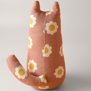 May include: A handmade stuffed cat toy in a terracotta color, adorned with a repeating pattern of white daisies with yellow centers. The cat has upright ears and a curved tail, all made from the same fabric.