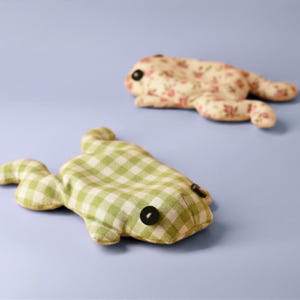 May include: Two handmade frog-shaped plush toys. One is green and white checkered, the other is cream-colored with a pink floral pattern. Both have black button eyes and are lying on a light blue surface.
