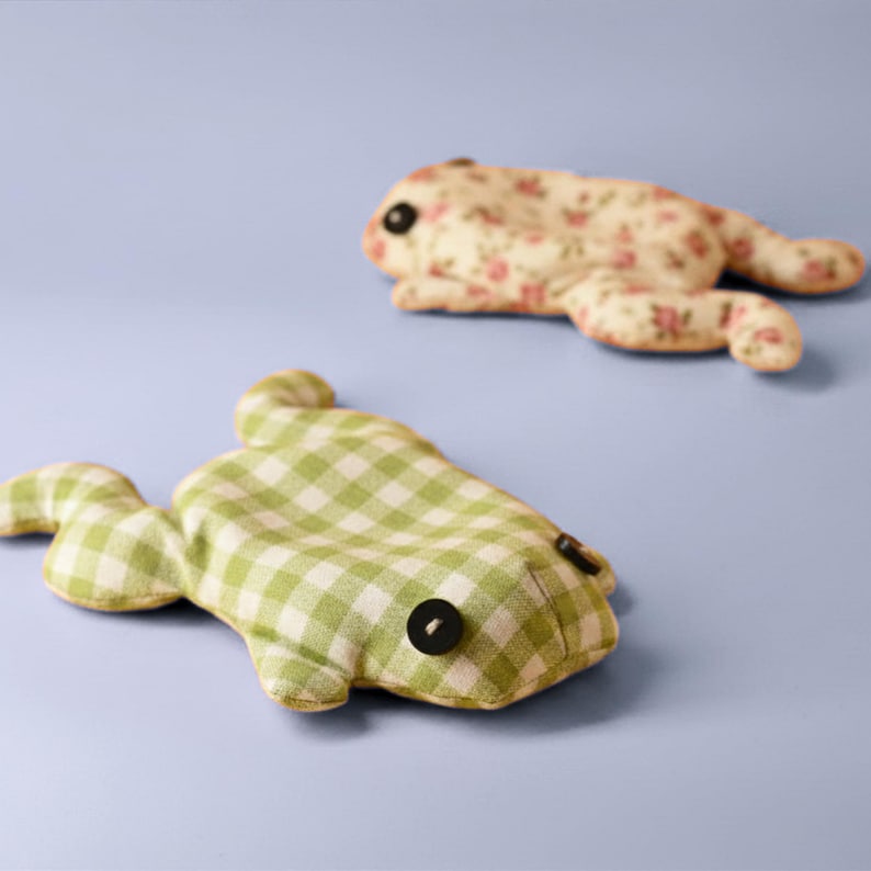 May include: Two handmade frog-shaped plush toys. One is green and white checkered, the other is cream-colored with a pink floral pattern. Both have black button eyes and are lying on a light blue surface.