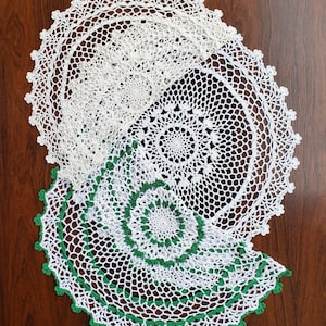 May include: White and green crocheted doilies arranged on a dark wood surface. The doilies feature intricate openwork patterns and scalloped edges. The green accents add a pop of color to the otherwise neutral palette, creating a vintage aesthetic.