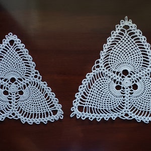 May include: Two white crocheted triangular doilies with intricate designs. The doilies have a delicate, lacy appearance and feature a repeating pattern of circles and swirls.