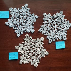 May include: Three handmade crochet snowflake doilies in white, cream, and silver. Each doily has a unique openwork design. The image includes sticky notes with the colors of the doilies: "White w/ Silver", "White w/ gold", and "Cream/off white w/ gold".