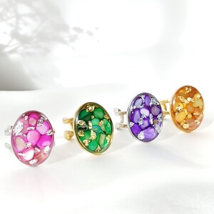 Pode incluir: Quatro anéis ajustáveis com engastes de pedras ovais coloridas. Os anéis apresentam pedras rosa, verdes, roxas e laranja com bandas douradas ou prateadas. As pedras têm uma aparência de mosaico com detalhes em folha de ouro.