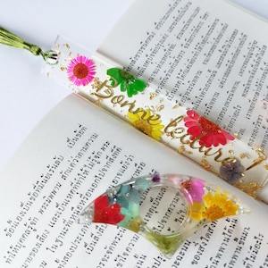 Può includere: Un segnalibro trasparente con fiori pressati e dettagli in foglia d'oro. Il segnalibro ha il testo "Bonne lecture" in lettere dorate.