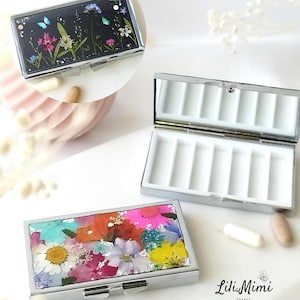 May include: Silver rectangular pill box with a hinged lid. One box features a black background with pressed flowers and butterflies. Another box has a white interior with seven compartments. A third box has a colorful floral design. The logo "Lili Mimi FRANCE" is visible.
