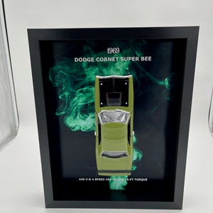 May include: A framed image of a green 1969 Dodge Coronet Super Bee model car. The car is shown from above with a green smoke effect behind it. The text "1969 Dodge Coronet Super Bee" and "440 V-8 4 Speed 390 HP 490 LB-FT TORQUE" is printed below the car.