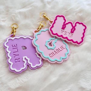 Peut inclure: Trois porte-clés lettres personnalisés avec des fermoirs mousquetons dorés. La lettre R violette porte le nom RYLIE, la lettre G rose porte le nom CHARLIE avec une bordure bleu clair, et la lettre M rose porte le nom ELSA avec une bordure rose foncé.
