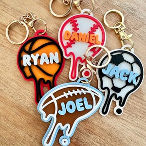 Puede incluir: Cinco llaveros personalizados con herrajes dorados. Los llaveros presentan diseños de balones deportivos: baloncesto, béisbol, balón de fútbol y fútbol americano. Cada llavero tiene un nombre en negrita: Ryan, Daniel, Jack y Joel. Los llaveros tienen un efecto de pintura goteando.