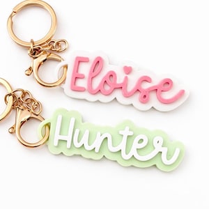 May include: Two personalised keyrings with gold-coloured hardware. One keyring features the name "Eloise" in pink script on a white background, with a small heart. The other has the name "Hunter" in white script on a light green background.