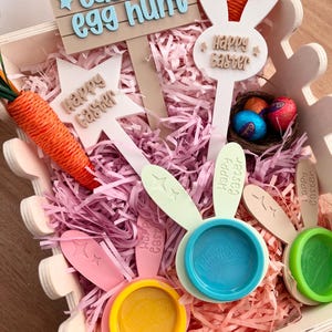 May include: A wooden Easter-themed box filled with pink paper shreds, decorative signs, and colorful Play-Doh containers. The signs read "Easter egg hunt" and "Happy Easter." A carrot and chocolate eggs are also included.