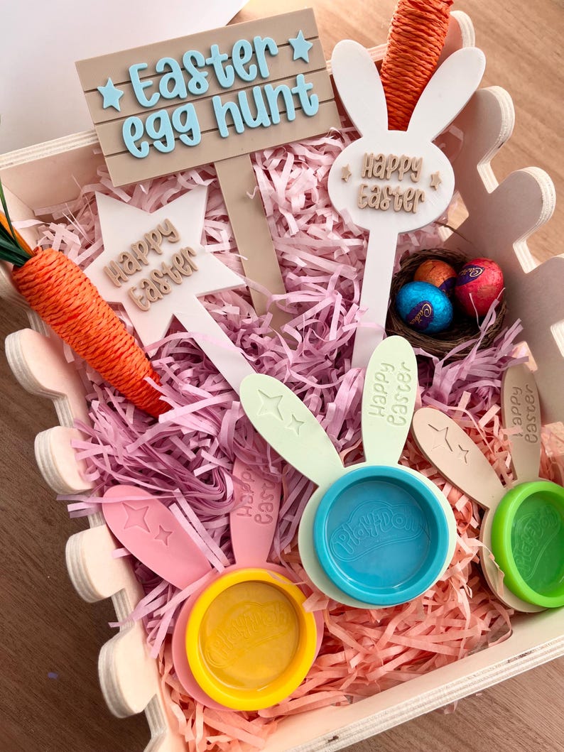 May include: A wooden Easter-themed box filled with pink paper shreds, decorative signs, and colorful Play-Doh containers. The signs read "Easter egg hunt" and "Happy Easter." A carrot and chocolate eggs are also included.