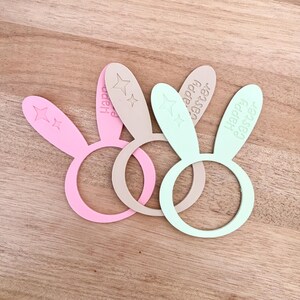 May include: Three pastel-colored Easter bunny ear napkin rings. The rings are pink, beige, and mint green. Each ear has a star design and the words "Happy Easter" engraved on them. The rings are on a wooden surface.