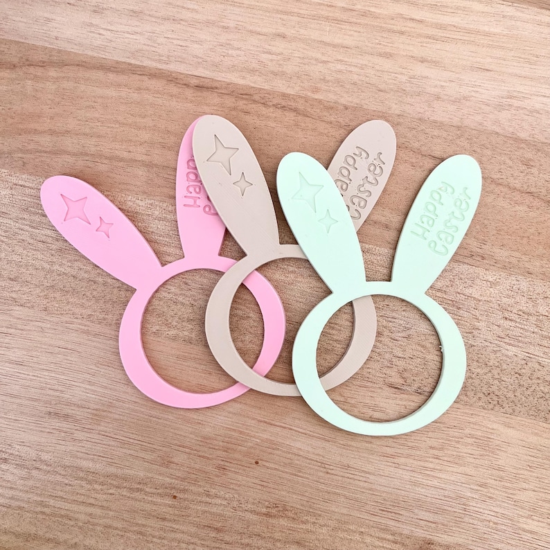 May include: Three pastel-colored Easter bunny ear napkin rings. The rings are pink, beige, and mint green. Each ear has a star design and the words "Happy Easter" engraved on them. The rings are on a wooden surface.