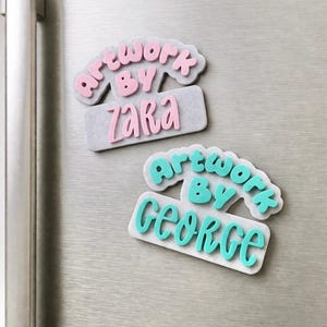 May include: Two decorative magnets with the text "Artwork by Zara" and "Artwork by George". The magnets are light grey with pink and teal lettering. The magnets are on a silver fridge door.