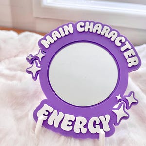 May include: A purple compact mirror with the words "MAIN CHARACTER ENERGY" in white lettering. The mirror is round and surrounded by a purple frame with star accents. The mirror is propped up by two white stands.
