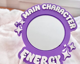 May include: A purple compact mirror with the words "MAIN CHARACTER ENERGY" in white lettering. The mirror is round and surrounded by a purple frame with star accents. The mirror is propped up by two white stands.