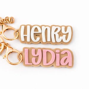 May include: Two personalized keychains with gold-colored clasps. One keychain reads "Henry" in white letters on a tan background. The other keychain reads "Lydia" in pink letters on a tan background. The keychains are likely made of acrylic.