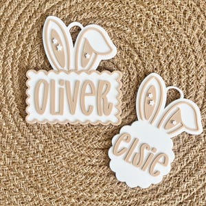 May include: Two Easter-themed name tags with bunny ears. One tag reads "Oliver" on a scalloped edge, the other reads "Elsie". The tags are white with beige accents and star details, set against a woven background.