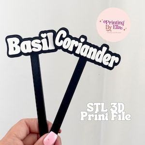 Op de afbeelding: Twee zwarte plantenmarkers met witte tekst die "Basilicum" en "Koriander" luidt. De tekst is zwart omlijnd. De afbeelding bevat ook de tekst "STL 3D Print File" en een roze cirkel met de tekst "Printing By Ella".