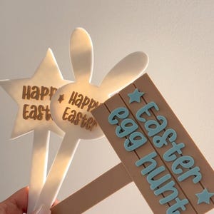 May include: Easter-themed yard stakes. The image shows three decorative stakes: a star, a bunny, and a sign that reads "Easter egg hunt." The stakes are white and brown with blue text. Perfect for Easter.