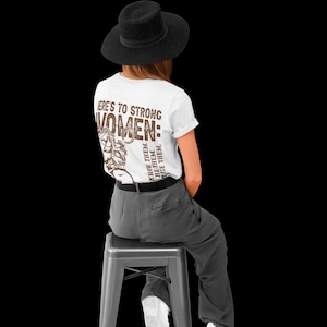 May include: A person wearing a white t-shirt with the text "HERE'S TO STRONG WOMEN" in brown. The shirt also features a graphic of a woman and the phrases "KNOW THEM. BE THEM. RAISE THEM."