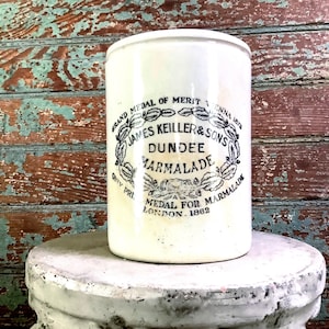 May include: A white ceramic jar with a cylindrical shape, featuring a black and white label that reads "JAMES KEILLER & SONS DUNDEE MARMALADE." The jar is set against a weathered wooden background, and is sitting on a white, round pedestal.