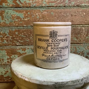 May include: A vintage, cylindrical ceramic jar with the text "FRANK COOPER'S OXFORD SEVILLE MARMALADE" in black lettering. The jar is off-white with a slightly aged appearance, sitting on a light green pedestal.