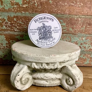 May include: A round white tin of Burgess's Genuine Anchovy Paste featuring a black printed crest and text including Hythe Road Willesden N.W and Late 107 Strand London. The tin is positioned on a weathered white stone pedestal with scroll details.