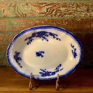May include: An oval, white ceramic dish with a scalloped blue rim and blue floral designs. The dish is supported by a gold-colored metal stand. The floral pattern is repeated around the dish's edge and center.