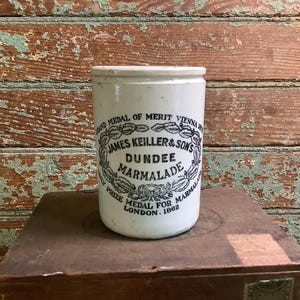 May include: A vintage white ceramic jar with a cylindrical shape. The jar features a black and white label with the text "JAMES KEILLER & SONS DUNDEE MARMALADE" and "PRIZE MEDAL FOR MARMALADE LONDON. 1862". The jar is placed on a wooden box.