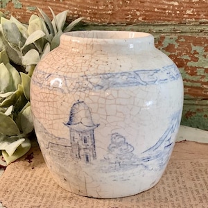 May include: A small, off-white ceramic vase with a crackle glaze. The vase features a blue landscape design with a building and trees. The vase has a round body and a wide opening, perfect for displaying small flowers or decorative items.