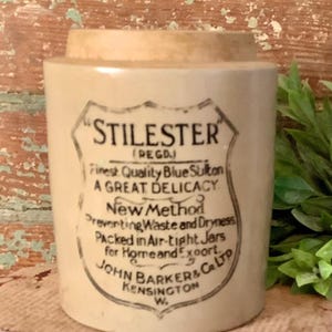 May include: A vintage stoneware jar with a beige body and a light brown lid. The jar features a black and white label with the text "STILESTER" and other details. The jar is cylindrical and appears to be an antique.