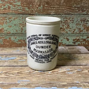 May include: Cream-colored antique marmalade jar with black text and a decorative emblem. The text reads "JAMES KEILLER & SONS DUNDEE MARMALADE" and "GRAND MEDAL OF MERIT VIENNA" with "LONDON. 1868" at the bottom. The jar is cylindrical with a slightly flared rim.