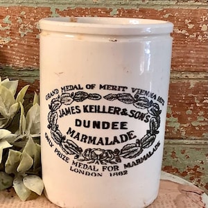 May include: A vintage white ceramic marmalade jar with black text and a decorative border. The text reads "JAMES KEILLER & SONS DUNDEE MARMALADE" and includes "GRAND MEDAL OF MERIT VIENNA 1870" and "ONLY PRIZE MEDAL FOR MARMALADE LONDON 1862".
