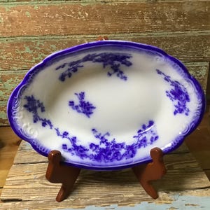 May include: An oval, white ceramic dish with a blue floral pattern and a scalloped blue rim. The dish is displayed on a small, brown wooden stand, set against a rustic wooden background.