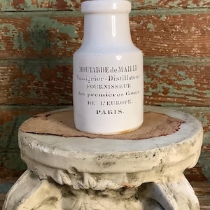 May include: A white ceramic jar with black text, labeled "Moutarde de Maille" and other French text. The jar sits on a decorative, off-white pedestal with a round top and scrollwork. The background is a distressed, multi-colored wooden surface.