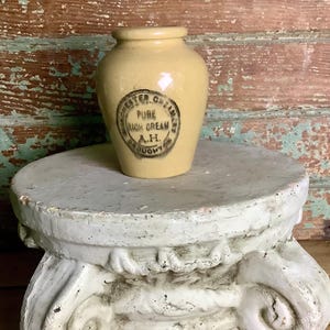May include: A small, light yellow ceramic jar with a rounded body and a short neck. The front features a circular black stamp with the text "CHESTER CREAMERY PURE RICH CREAM A.H. BROUGHTON" printed in the center of the vessel's smooth surface.