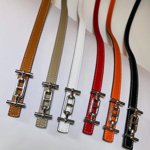 May include: A selection of leather straps in shades of brown, beige, white, red, orange, and black. Each strap is fitted with a silver-toned metal clasp. The straps are displayed in a line, highlighting their diverse colours and textures.