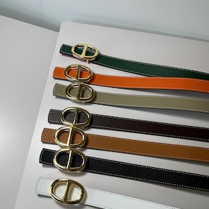 Puede incluir: Una colección de siete cinturones en varios colores, incluyendo verde, naranja, beige, marrón oscuro, tostado, negro y blanco. Cada cinturón presenta una correa texturizada con costuras blancas en contraste y una hebilla ovalada dorada pulida.