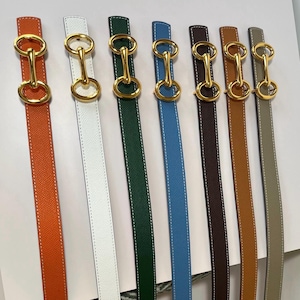 May include: Seven leather belts in orange, white, dark green, blue, dark brown, tan, and grey, each featuring a gold-tone metal buckle with a central horizontal bar connecting two circular rings, finished with white contrast stitching along the edges.