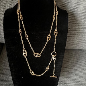 Puede incluir: Collar dorado con diseño de doble cadena. El collar presenta eslabones ovalados y un cierre de barra. La joya se exhibe en un maniquí negro, mostrando su elegante diseño y sus intrincados detalles.