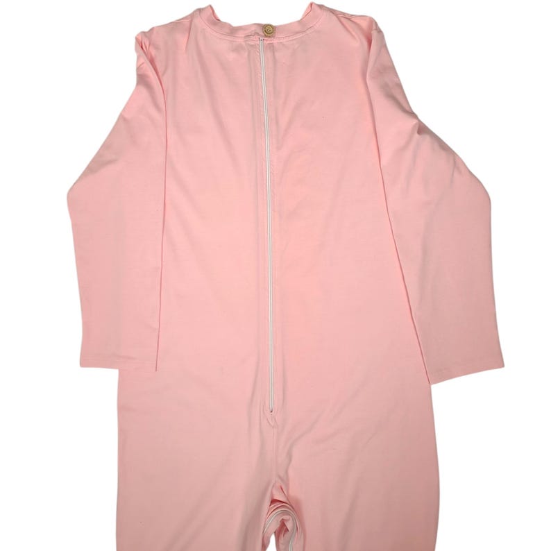 Pode incluir: Um macac&atilde;o de pe&ccedil;a &uacute;nica em rosa claro liso, com mangas compridas, decote redondo com um pequeno fecho de bot&atilde;o no topo e um fecho de correr branco vertical que percorre o centro da parte frontal do tronco.