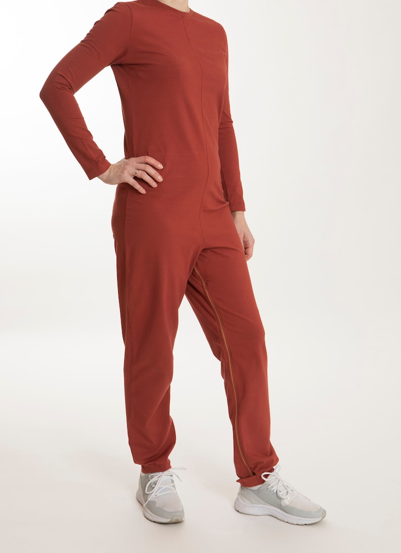 Pode incluir: Um macac&atilde;o de manga comprida e comprimento total numa cor vermelho terracota s&oacute;lido. A pe&ccedil;a apresenta um decote redondo, uma costura vertical ao longo do centro do tronco e um fecho de correr longo que vai da virilha at&eacute; &agrave; perna.