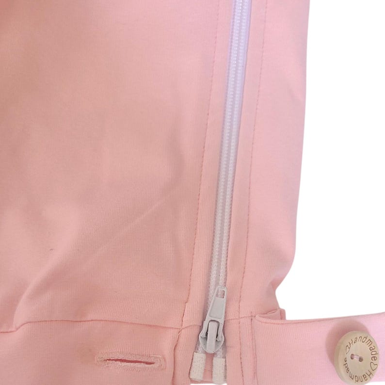 Pode incluir: Uma pe&ccedil;a de vestu&aacute;rio em tecido rosa claro com um fecho de correr vertical branco. Um pequeno bot&atilde;o de madeira redondo e claro est&aacute; fixado no lado inferior direito, gravado com o texto "Handmade" e um pequeno s&iacute;mbolo de cora&ccedil;&atilde;o.
