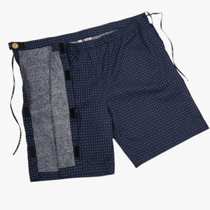 May include: Navy blue shorts with a white polka dot pattern. The shorts feature a button closure and a contrasting grey panel with velcro fastenings. Drawstrings are visible at the waist.