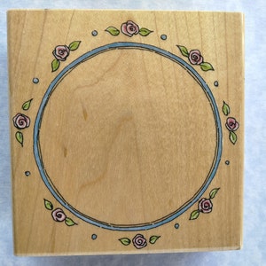 May include: A wooden stamp with a blue and white circle border decorated with pink roses and green leaves. The circle is empty for stamping.