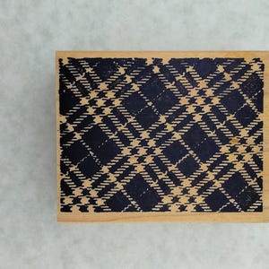May include: A rectangular wooden stamp with a navy blue and tan plaid pattern. The stamp has a detailed, diagonal design. The wood has a light brown color. The stamp is on a white surface.