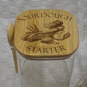 May include: A clear glass jar with a wooden lid. The lid has the words "Sourdough Starter" engraved on it, along with a design of bread loaves and wheat.