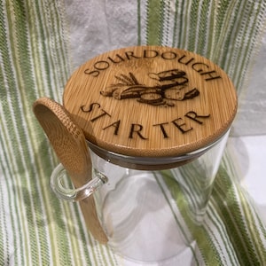 May include: A glass jar with a bamboo lid and a wooden spoon. The lid has the words "Sourdough Starter" engraved on it.